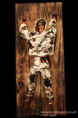 N°87: L'homme à la capuche
						Création 2026
						Dimensions: 42X18X8 cm. 
						Auteur; Loïc Jugue
						Technique : Poupée Action man avec une capuche, brûlée et clouée sur du bois de récupération. 