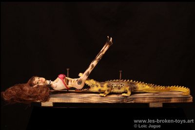 N°88: La blonde et le crocodile
						Création 2026
						Dimensions: 49X15X15 cm. 
						Auteur; Loïc Jugue
						Technique : Poupée femme (Barbie) et crocodile en plastique tous les deux brûlés et cloués sur une planche de palette brûlée. 