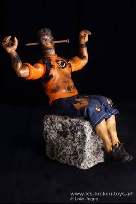 N°92: Les assis 1 
						Création 2026
						Dimensions: 24X17X16 cm. 
						Auteur; Loïc Jugue
						Technique :Poupée homme, lunettes noires, avec un clou dans la tête, assis (collée) sur un bloc de granit.  