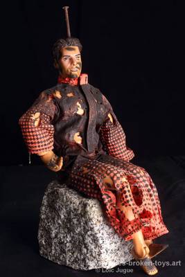 N°93: Les assis 2
						Dimensions: 24X17X16 cm. 
						Auteur; Loïc Jugue
						Technique :Poupée homme, avec un clou dans la tête, il porte une combinaison orange avec des points noirs. Il assis (collée) sur un bloc de granit.   