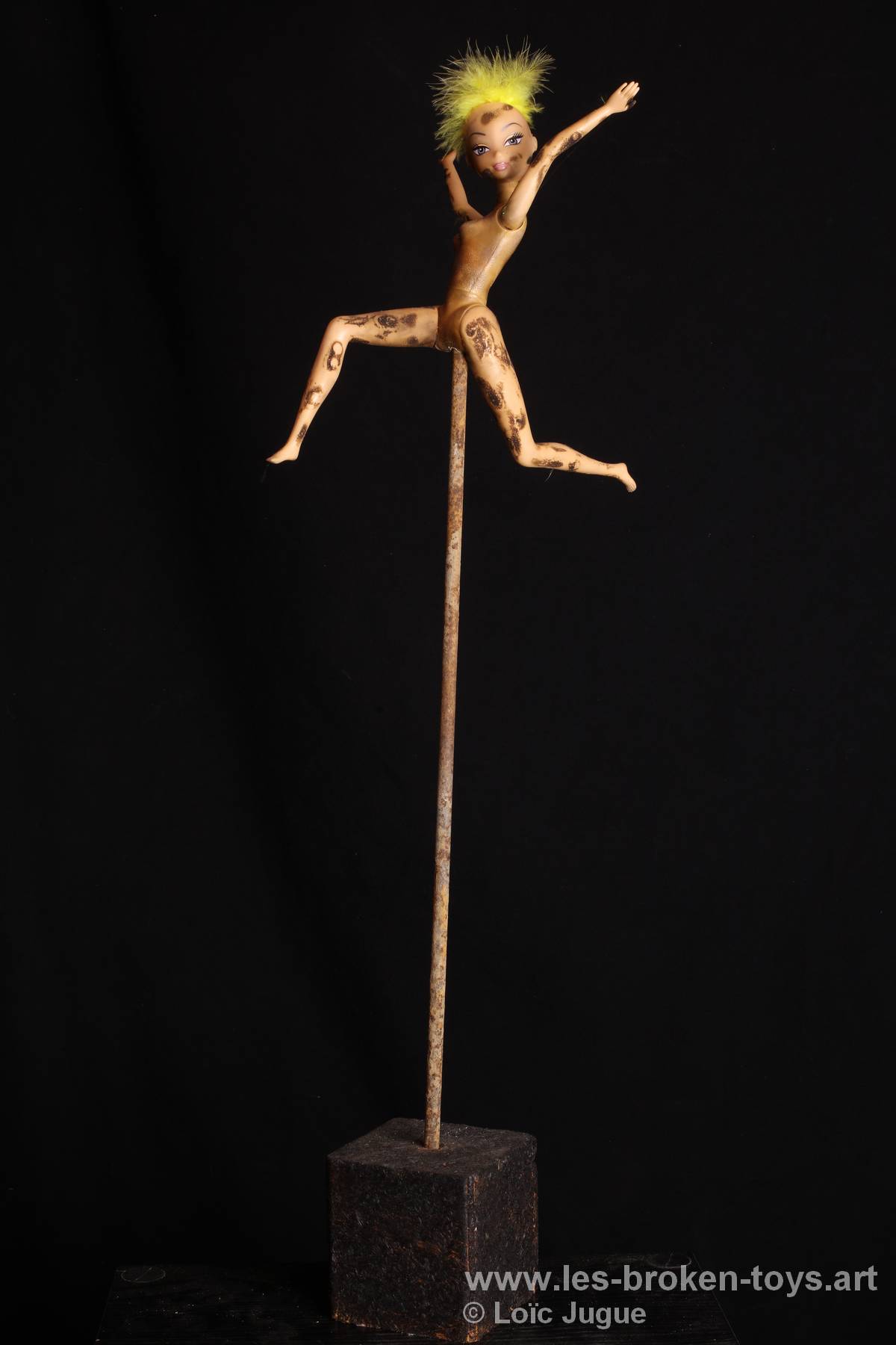 N°96: Coureuse à la chevelure jaune
						Dimensions: 72X20X26 cm. 
						Auteur; Loïc Jugue
						Technique :Poupée nue brûlée, plume jaune, barre en métal, cube en bois de récupération.