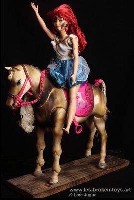 N°99: La cavalière rousse
						Création 2026
						Dimensions: 39X30X10 cm. 
						Auteur; Loïc Jugue
						Technique :Poupée rousse, robe bleue, brûlée. Cheval à la selle rose. Socle en bois de palette brûlé. 