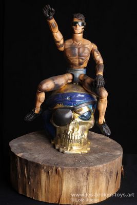 N°95: Jeff Koons chevauchant Damien Hirst, 
						Création 2023, 
						Dimensions: 40X27X16 cm. 
						Auteur; Loïc Jugue
						Technique : Poupée homme brûlée, lunettes noires,  tête de mort dorée, socle en bois.